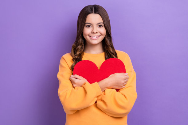 可爱开朗的女孩的照片，牙齿微笑，手臂抱红纸心脏符号孤立的紫色背景图片下载