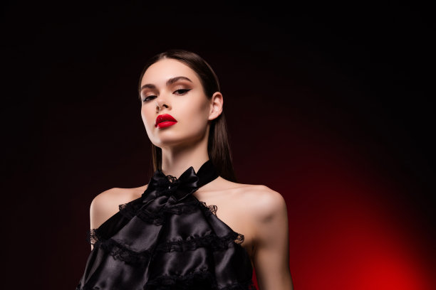 照片中的女士穿着黑色晚礼服，有着明亮的妆容，在相机中向下看，孤立的红色背景图片下载