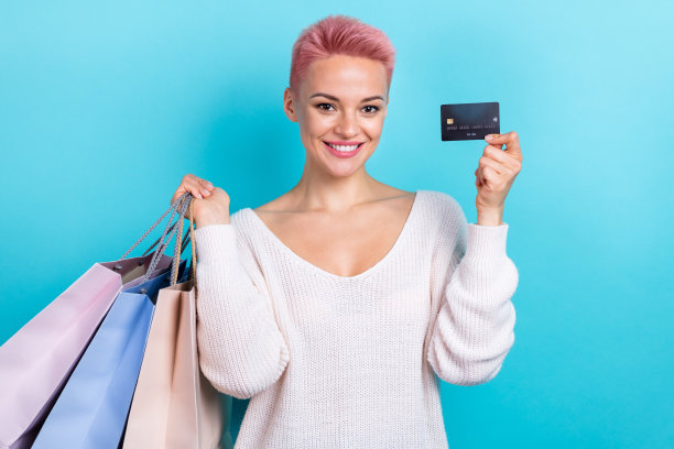 照片中年轻的粉色短发女士穿着时尚的蓝色长袖，手持塑料银行卡，在青色背景上花钱购物袋图片下载