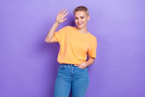 照片中的年轻友好的女孩穿着橙色t恤时尚牛仔裤，显示手掌hi五hello世界广告衣服孤立的紫色背景图片下载