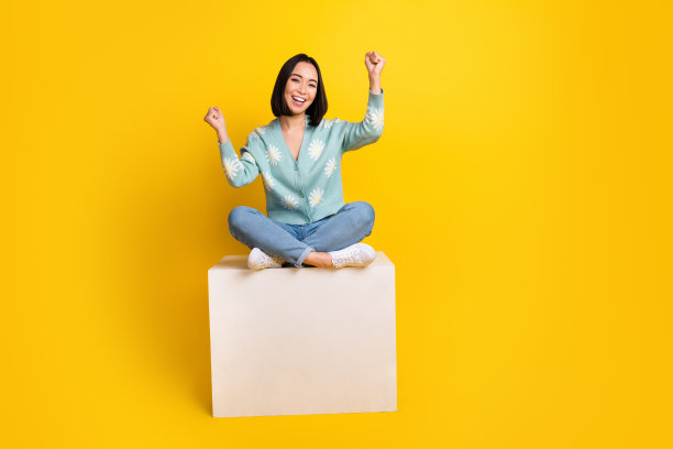 大喜大喜满意的女人的全尺寸照片蓝色套头衫坐在立方体上握紧拳头孤立的黄色背景图片下载