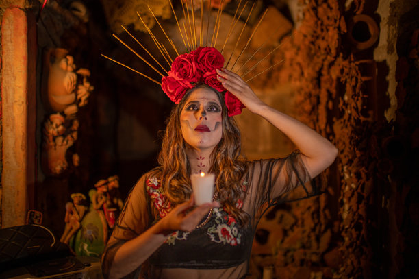 一个墨西哥女人打扮成卡特里娜，头上戴着红色的花朵头饰，在一个古老、黑暗的房子里拿着蜡烛，抬头看，在一张水平照片中。图片下载