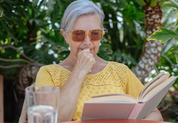 一位老妇人在户外看书、喝水，享受着宁静的时光。这张照片捕捉到了自然环境中的宁静、自我照顾和智力参与。图片下载