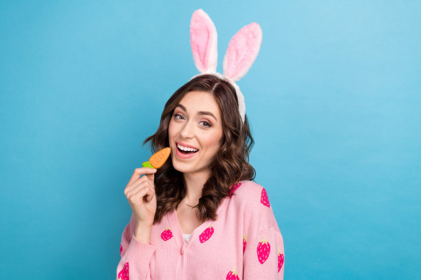 照片中的兴奋闪亮的女人穿着粉红色的开襟羊毛衫有发带咬胡萝卜形状饼干孤立的蓝色背景图片下载