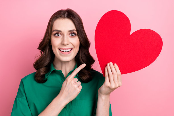 照片上光鲜亮丽的女子穿着绿色衬衫，用手指指着红色的心，空白的空间孤立出粉红色的背景图片下载