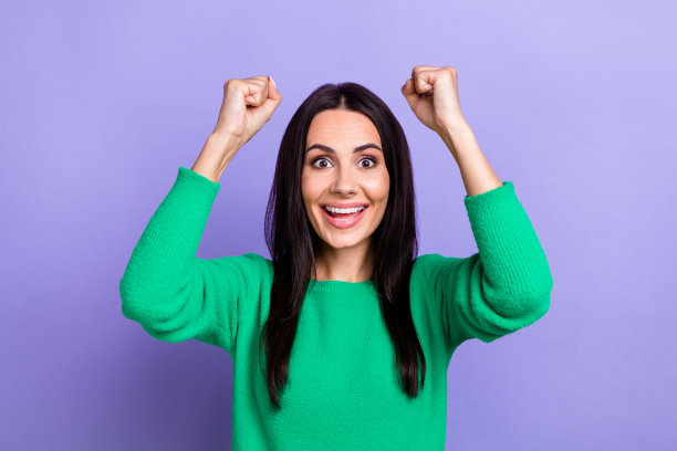 照片中漂亮幸运的女人穿着绿色套头衫，尖叫着举起拳头，孤立的紫色背景图片下载