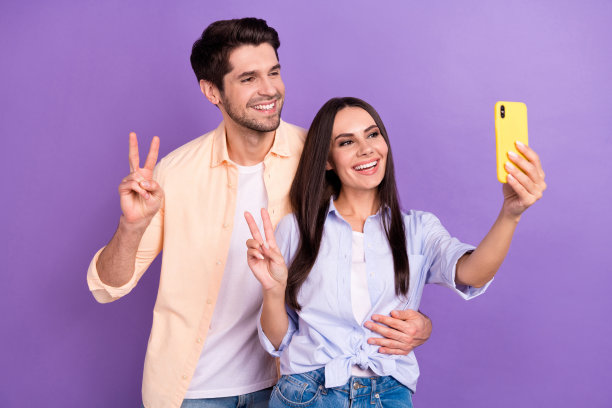 这是一对愉快友好的情侣情侣的自拍照片，他们展示了v-sign hello符号，他们的新智能手机视频被隔离在紫色背景上图片下载