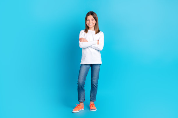 乐观愉快的女孩穿白色长袖牛仔裤站立双臂交叉孤立在蓝色背景的全尺寸照片图片下载
