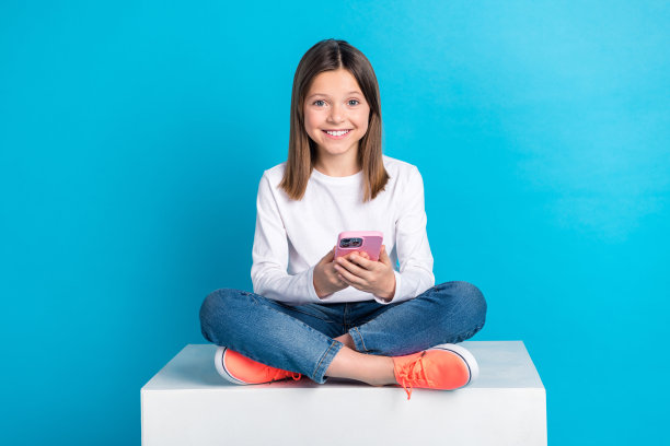 快乐的女孩穿白色长袖牛仔裤的全尺寸照片，阅读通知，坐在立方体上孤立的蓝色背景图片下载