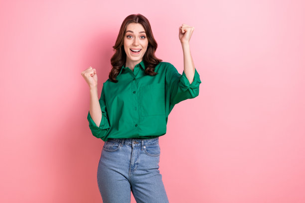 照片正面印象可爱的女人波浪发型穿着绿色衬衫举起拳头赢得孤立的粉红色背景打赌图片下载