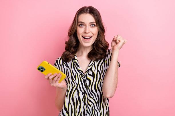 欣喜若狂的年轻女孩的照片举起拳头庆祝玩得开心玩电话在线赌博彩票棕色波浪的头发孤立在粉红色的背景图片下载