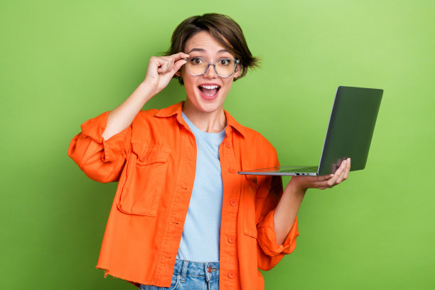 照片中，开朗可爱的女士穿着时髦的橙色衣服，拿着上网本设备，在绿色的背景下进行在线学习图片下载