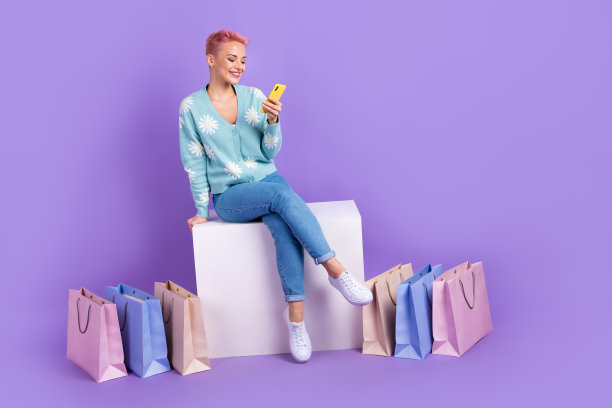 年轻女士的全身照片，穿着时髦时髦的衣服，用手机上网购物，坐在盒子里，选择衣服，孤立在紫罗兰色的背景上图片下载