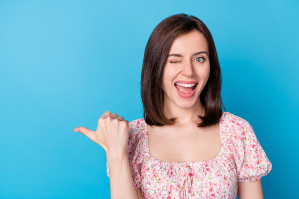 年轻漂亮女子的特写照片，眨眼的眼睛指导手指空白空间推荐新产品隔离在蓝色背景图片下载