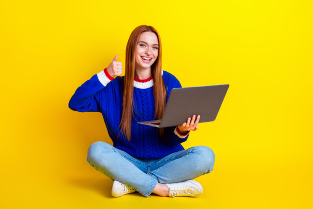 迷人的年轻女子的全身照片拿着上网本拇指朝上坐在地板上穿着蓝色的针织衣服孤立在黄色的背景图片下载