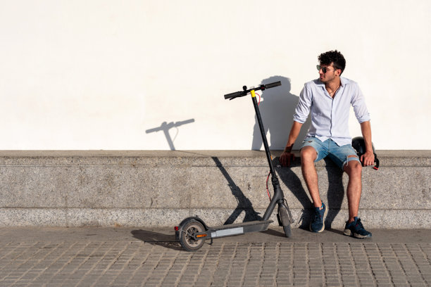 年轻人在一个阳光明媚的日子里骑着电动滑板车和头盔在墙上放松图片下载