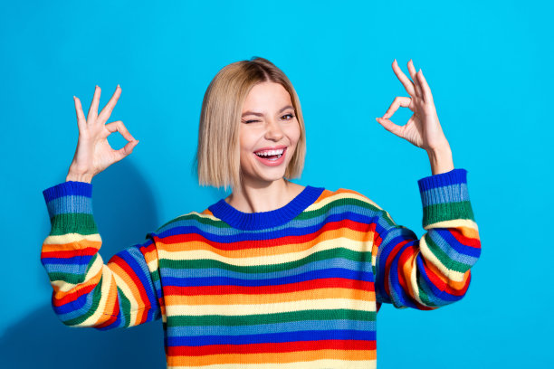 漂亮的年轻女子的照片，眼睛眨着眼睛，显示出好的标志，穿着条纹毛衣，孤立在蓝色背景上图片下载