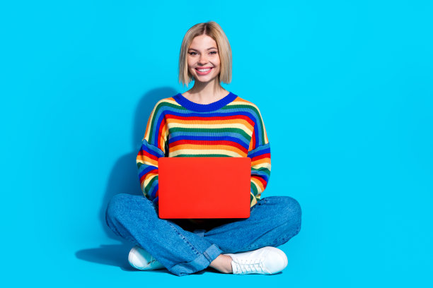 可爱的年轻女士坐在地板上拿着上网本的全身照片，穿着时尚的条纹服装，孤立在蓝色背景上图片下载