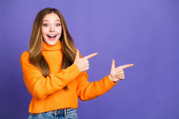 照片的青年迷人的金发可爱的快乐的微笑女孩在橙色高领毛衣指着手指广告牌孤立的紫罗兰色背景图片下载