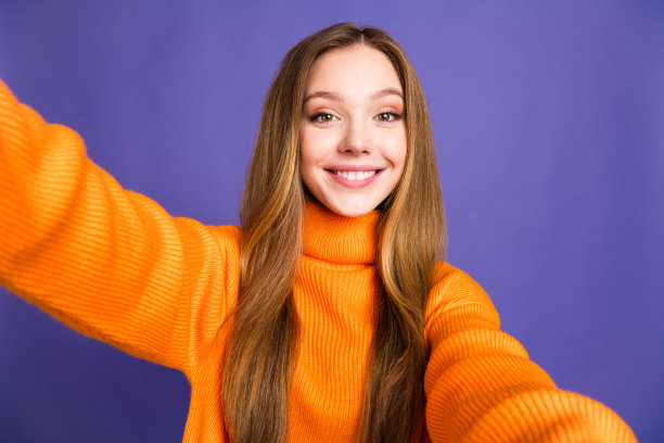 自拍照片的青年迷人的金发可爱的快乐的牙齿灿烂的微笑女孩在橙色毛衣孤立的紫罗兰色背景图片下载