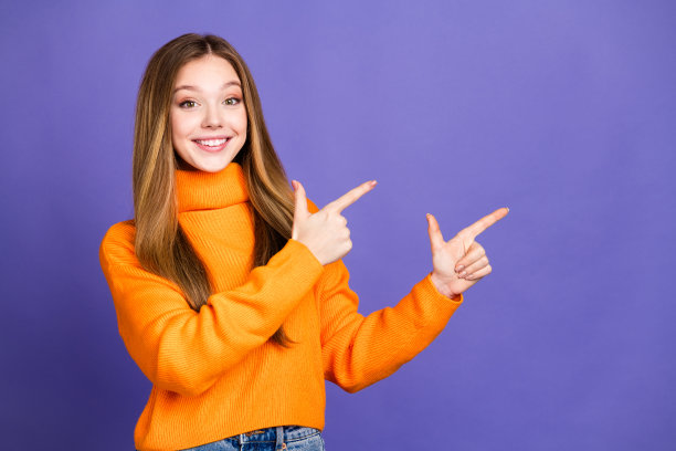 青春的肖像照片迷人的金发可爱的快乐的微笑女孩在橙色高领毛衣食指空白的空间孤立在紫色的背景图片下载