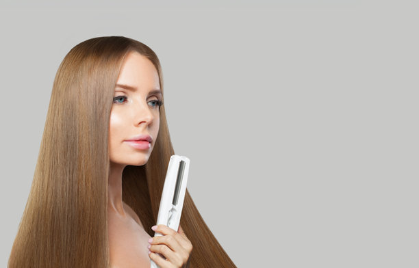 优雅的黑发女人拿着直发棒，在白色的背景上拉直她光滑闪亮的长发。护发、发型、美发及发型设计理念图片下载