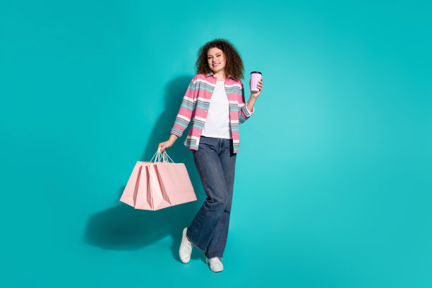 波浪头发的年轻女子在时尚的休闲服装拿着购物袋与最喜欢的咖啡孤立的海蓝宝石色背景的照片图片下载