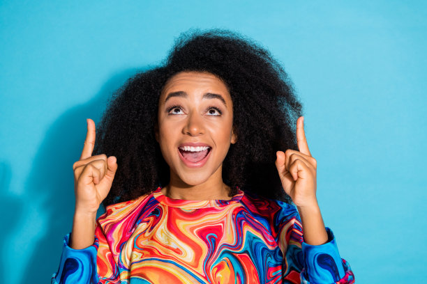 美丽的卷发惊讶的女人穿着时髦的运动衫手指指着头顶孤立的蓝色背景的照片肖像图片下载