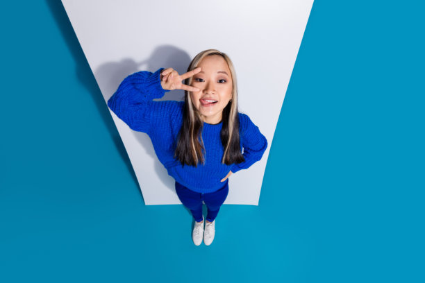 俯视图照片，时髦的韩国女孩靠近手机显示盖脸v标志摆姿势伸出舌头孤立的蓝色背景图片下载