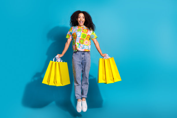 迷人的正面女孩穿着印花t恤跳跃抱着购物者空的空间孤立的蓝色背景的全长照片图片下载