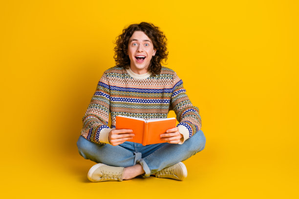 印象深刻的家伙的全尺寸照片穿着装饰毛衣坐在地板上阅读有趣的书孤立的黄色背景图片下载