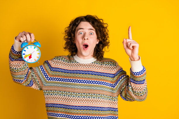 照片中聪明的小伙子有着波浪状的发型，穿着装饰毛衣，举起手指，在黄色的背景上拿着警报器图片下载