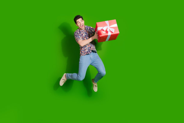 狂喜的家伙穿着印花衬衫牛仔裤的全尺寸照片，跳着拿着礼物看着销售空白的空间孤立在绿色的背景上图片下载