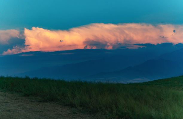 云是一个笑脸。从长满绿草的小山上看到一片明亮的大云，被夕阳的光线染成颜色，远处是高山。云朵的右边露出了一丝微笑。水平的照片。图片下载