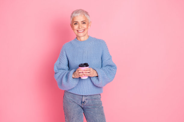 漂亮的退休女性的照片肖像拿着咖啡纸杯，穿着时髦的蓝色针织服装，孤立在粉红色的背景上图片下载
