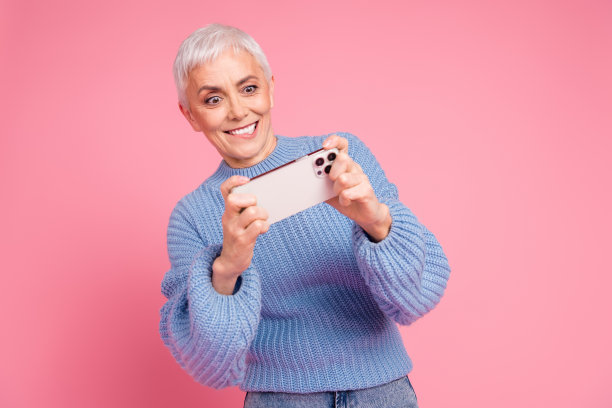 漂亮的退休女性玩手机游戏的照片肖像穿着时髦的蓝色针织服装孤立在粉红色的背景图片下载