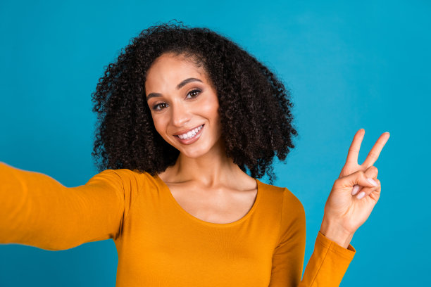 漂亮的年轻女孩的照片肖像自拍照片显示v字穿着时髦的黄色衣服孤立的蓝色背景图片下载