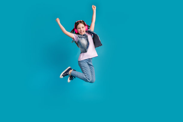 迷人的小女孩的全尺寸照片耳机跳举起拳头赢得穿时髦的牛仔裤服装孤立的蓝色背景图片下载