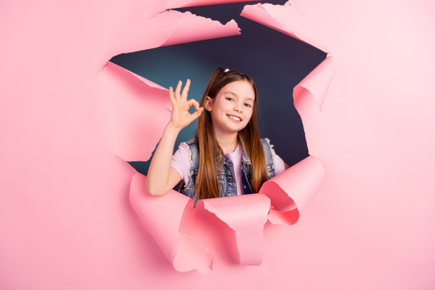 可爱的小女孩的照片肖像显示ok撕开的纸海报穿着时尚的牛仔服装孤立的蓝色背景图片下载