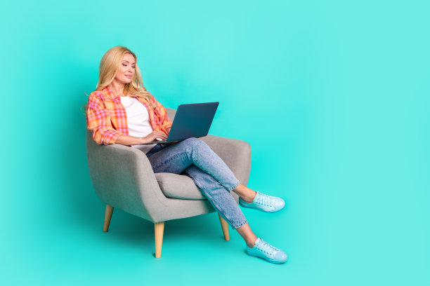 迷人的成熟的女人的全身照片，坐在扶手椅上，拿着上网本，穿着时尚的格纹衣服，孤立在青色背景上图片下载