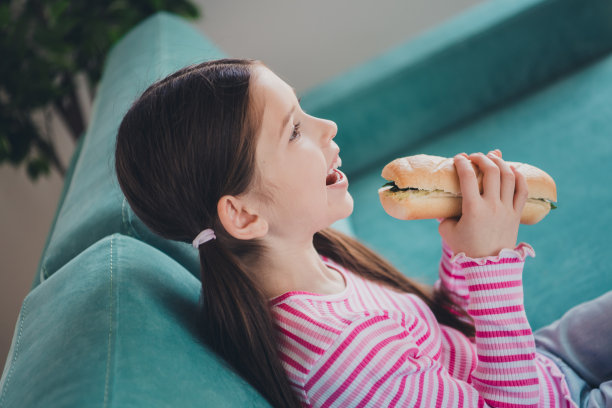 照片肖像的年轻有趣的孩子小黑发女孩呆在家里慢动作咬法棍三明治在长沙发上孤立在她的房间图片下载