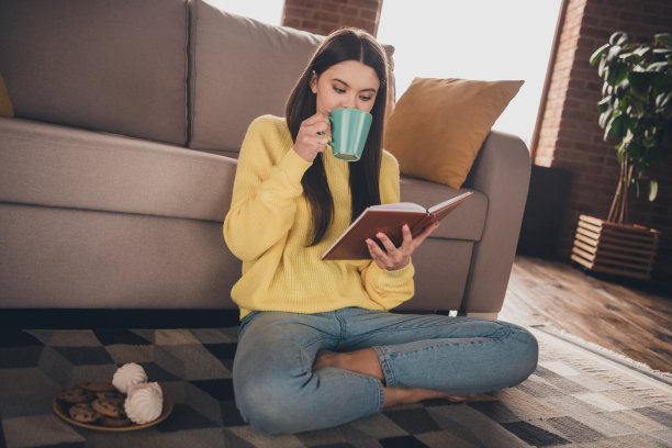 迷人少女的全尺寸照片坐在地板上喝茶零食看书穿着黄色的衣服舒适的日光家室内客厅图片下载