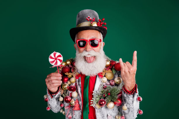 酷酷的退休老人圣诞老人的照片男性摇滚符号持有糖果打扮的球小玩意在夹克帽子太阳镜孤立的绿色背景图片下载