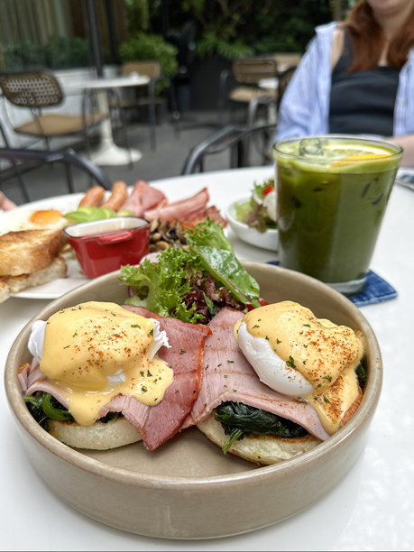 碗里装着本尼迪克特鸡蛋和牛油果、菠菜和猕猴桃汁混合的绿色饮料，冰块，放在牛仔布杯垫上，碗里装着生菜、樱桃番茄和萝卜片的配菜沙拉，白色盘子里的油炸早餐，露天用餐的女人图片下载