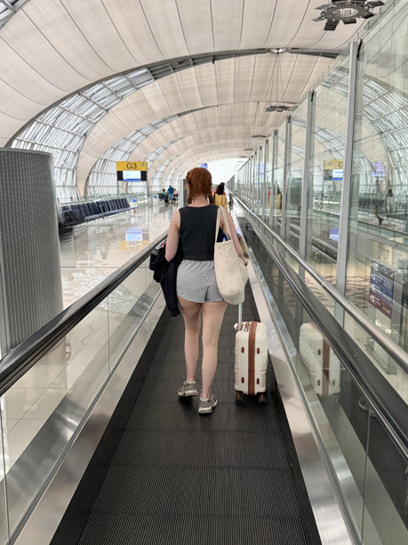 照片中，红头发的女旅客穿着休闲服装、短上衣、短裤和运动鞋，带着飞机行李、手提袋，乘坐电梯旅行，带伸缩把手的轮式行李箱，摄于机场航站楼，后视图，焦点在前景图片下载