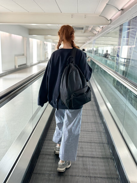 红发女旅客的特写照片，穿着休闲服装，海军蓝运动衫，条纹阔腿裤和运动鞋，背上有帆布袋，在机场候机楼的电梯上旅行，后视图，焦点在前景图片下载