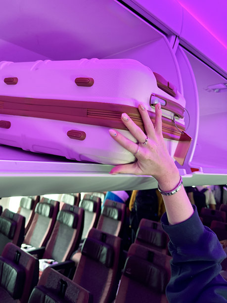 特写照片中，一个无法辨认的女人把带轮子的行李箱放在飞机机舱上方的行李舱里，粉红色的霓虹灯照亮了舒缓的登机心情，一排座位，聚焦在前景上图片下载