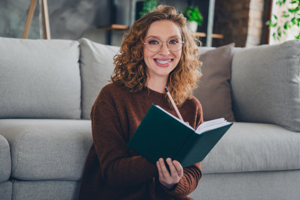 开朗迷人的女士的照片，穿着时髦的毛衣，坐在地板上，拿着笔记本，本，在家里写笔记图片下载