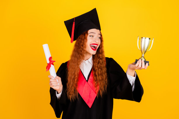 可爱的开朗的女孩的照片穿着黑色的斗篷，拿着奖杯和文凭孤立在黄色的背景图片下载