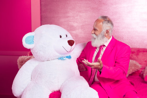 迷人的成熟的祖父男子的照片坐在沙发上和白色的大玩具熊说话穿粉红色的夹克在房间内部图片下载